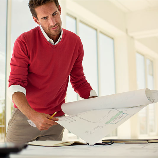 A person wearing a red sweater leans over a desk while reviewing large architectural blueprints