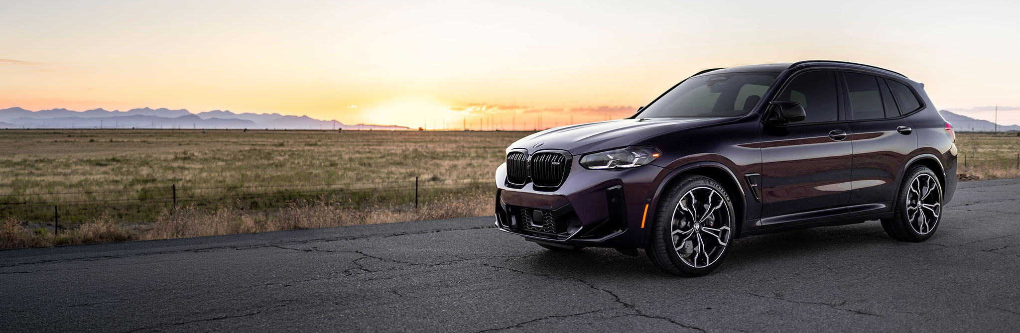 A dark-colored performance SUV parked on a cracked desert road at sunset, with mountains silhouetted on the horizon. 