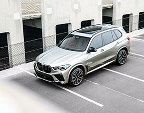 A silver performance SUV parked in an empty parking lot beside a concrete building with open garage bays and chain-link fencing.