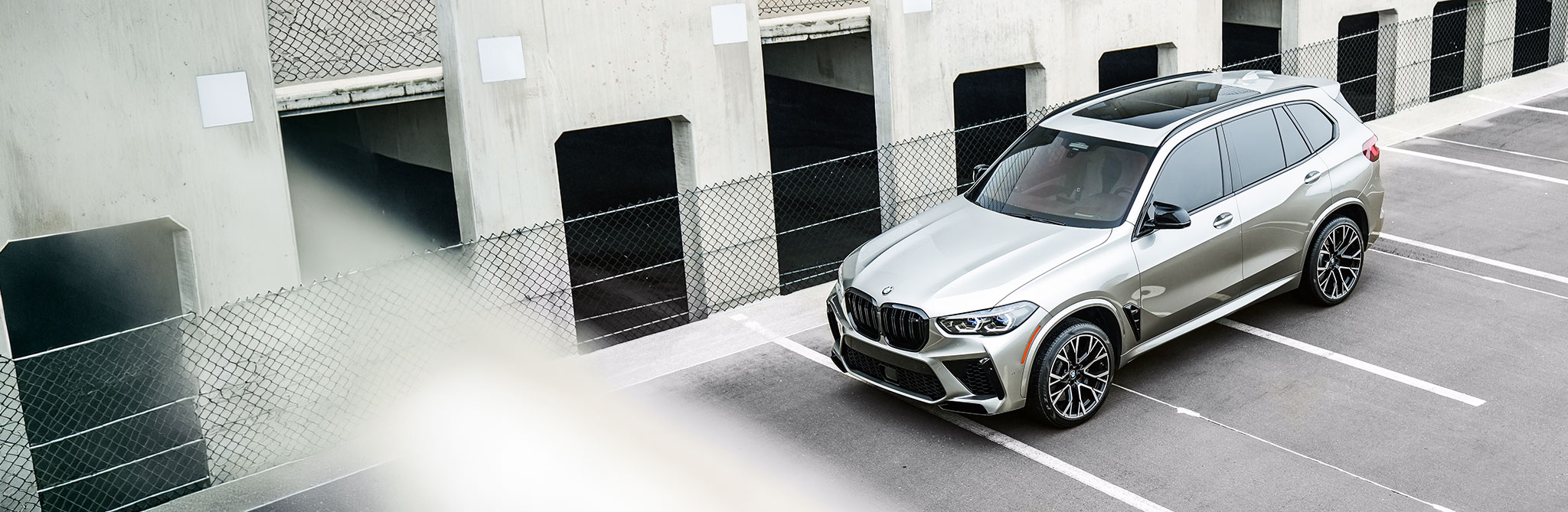 A silver performance SUV parked in an empty parking lot beside a concrete building with open garage bays and chain-link fencing. 