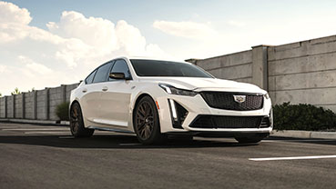 A white sports sedan parked on a wide roadway next to a long concrete barrier, with trees and a cloudy sky in the background.