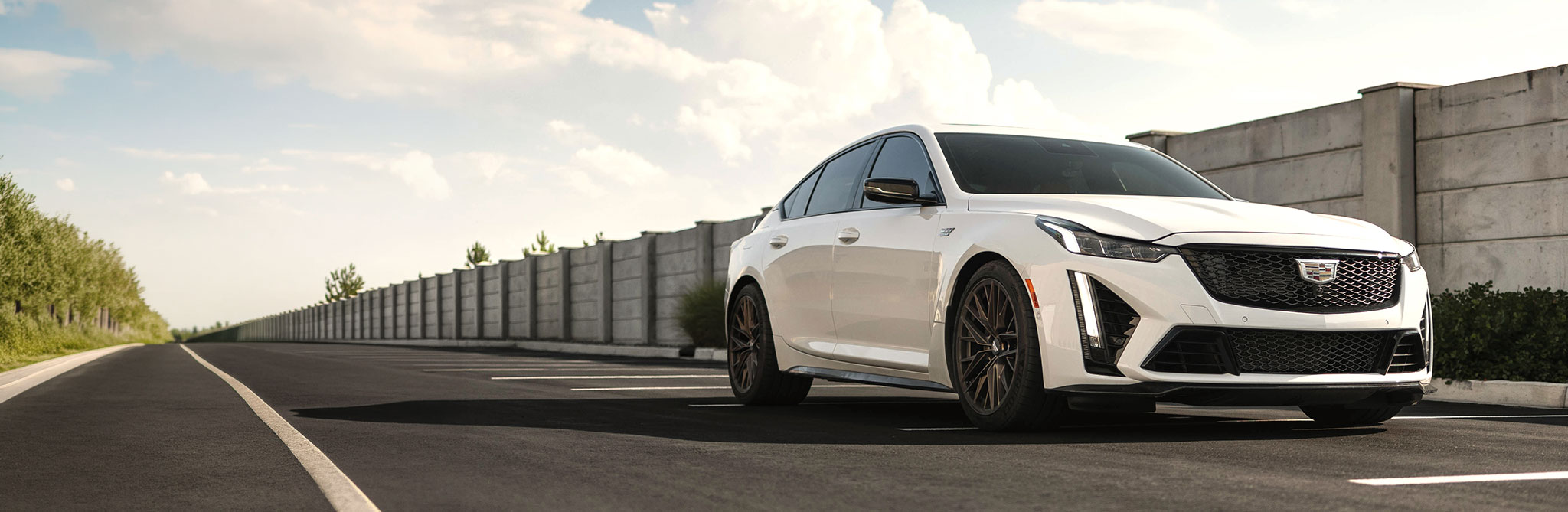 A white sports sedan parked on a wide roadway next to a long concrete barrier, with trees and a cloudy sky in the background. 