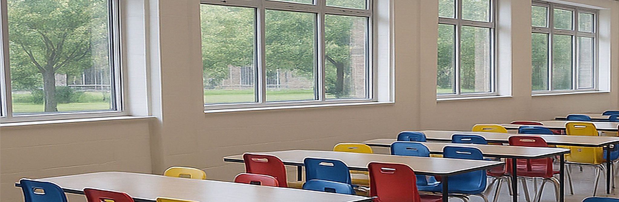 Bright cafeteria with long tables and colorful chairs next to large windows overlooking trees. 