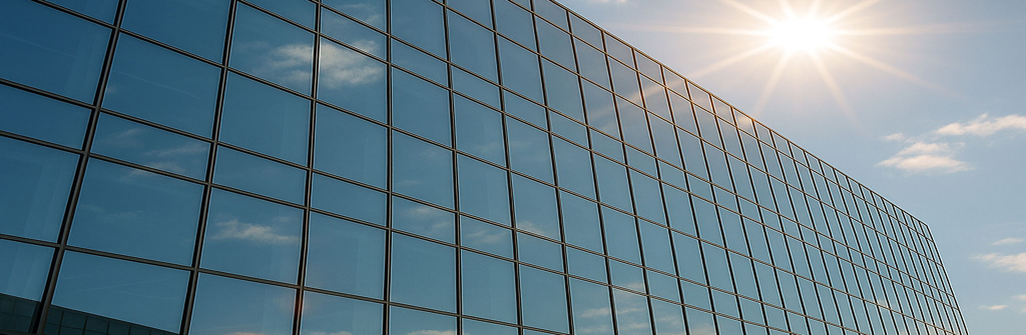 A modern glass office building reflecting the blue sky and bright sun, showcasing large architectural window panels. 