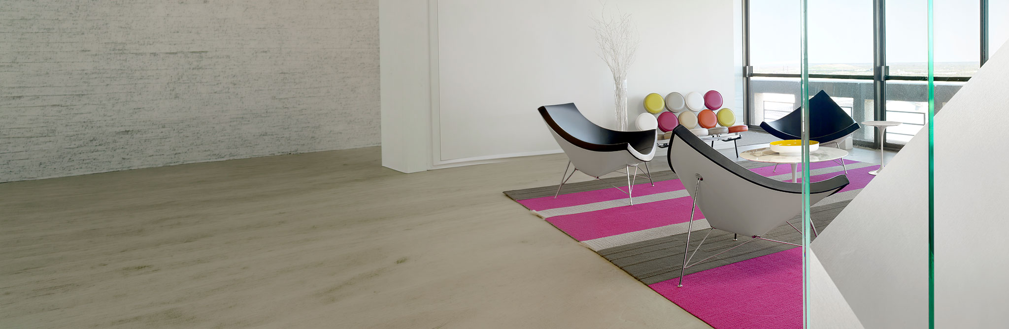 A contemporary interior space with minimalist furniture, including black lounge chairs, a colorful rug, and large floor‑to‑ceiling windows overlooking an outdoor view. 