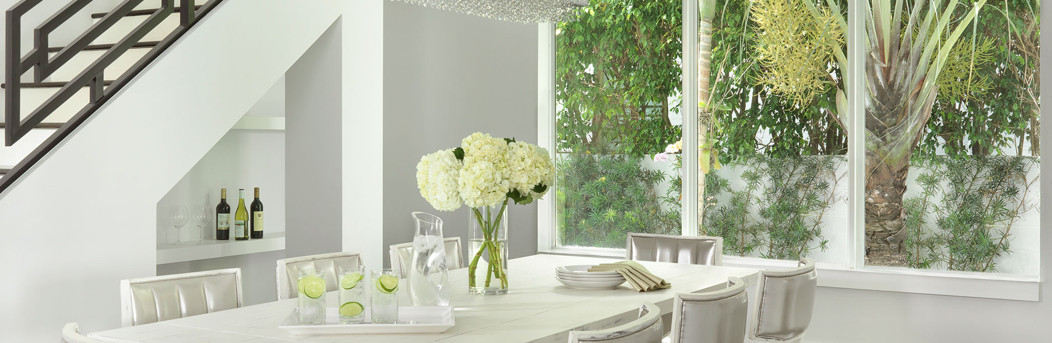 Bright dining room with a white table, neutral chairs, and large windows overlooking lush greenery. 