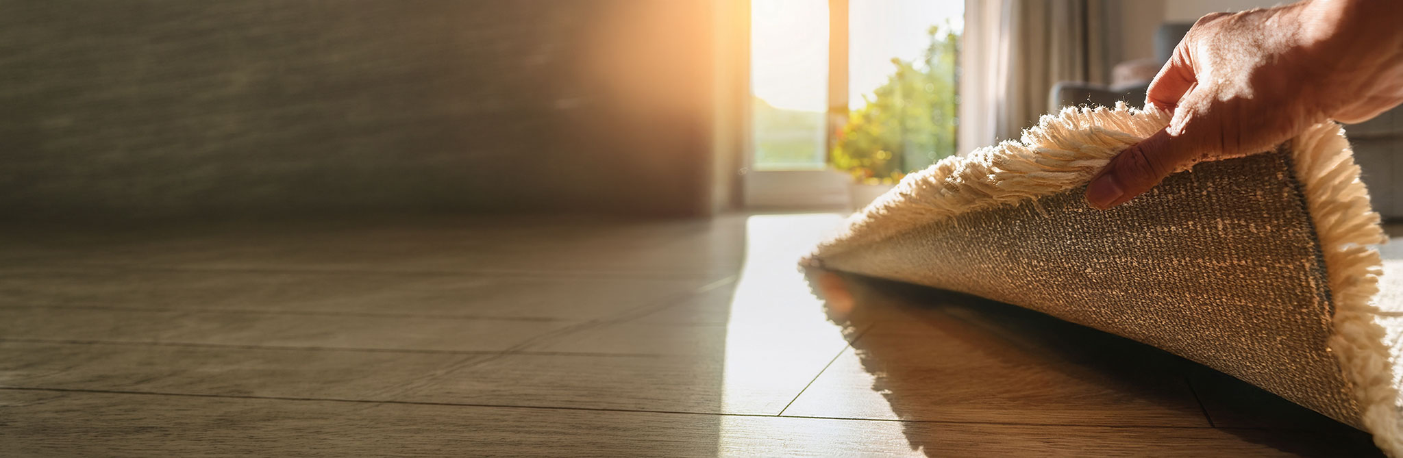 A hand lifting the edge of a textured rug to reveal sunlight shining across a hardwood floor near a large window. 