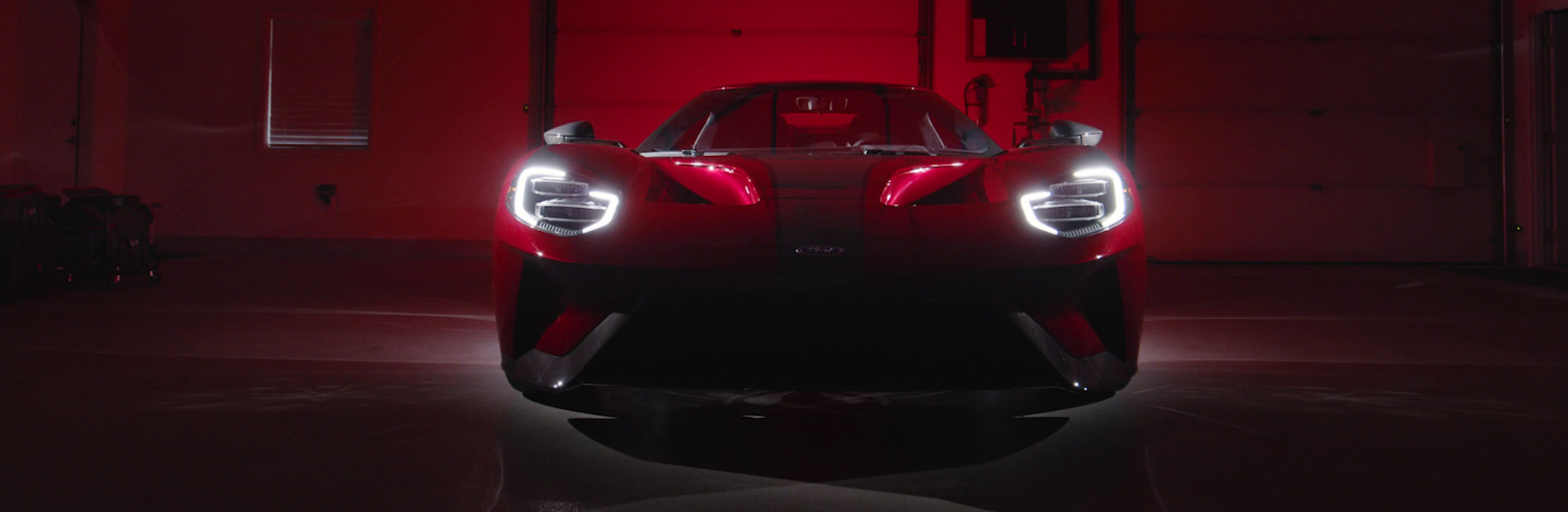 A low-lit view of a red sports car facing forward in a dark garage, with its bright LED headlights glowing against the red background. 