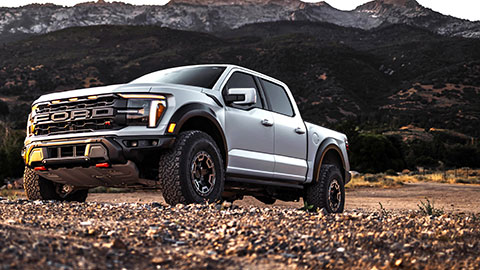 A silver off‑road pickup truck parked on rocky terrain with tall mountain peaks and evening light in the background.
