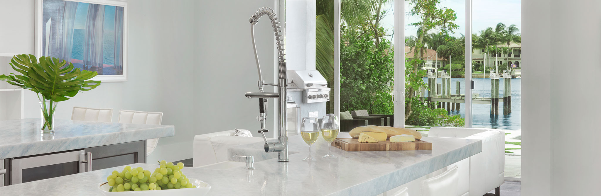 A bright modern kitchen with a large marble island, chrome pendant lights, white seating, and floor‑to‑ceiling windows overlooking lush greenery and a waterfront dock. 