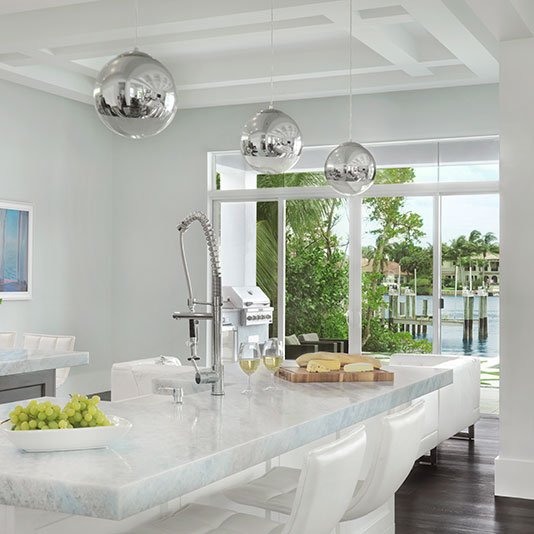 A bright, modern kitchen with white cabinetry and a large marble island, featuring chrome pendant lights, a tall spring faucet, and large windows overlooking lush greenery and a waterfront view.