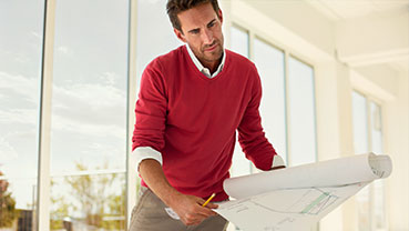 A person wearing a red sweater leans over a desk while reviewing large architectural blueprints