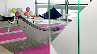 A contemporary interior space with minimalist furniture, including black lounge chairs, a colorful rug, and large floor‑to‑ceiling windows overlooking an outdoor view.