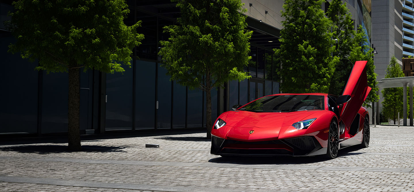 Red exotic sports car with scissor door open parked on a modern city street lined with trees. 