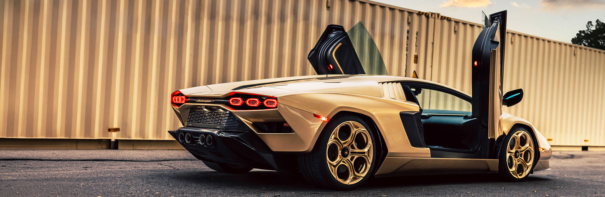 A gold-colored supercar parked on pavement with both scissor doors open, shown from a rear three‑quarter angle, with tall shipping containers in the background. 