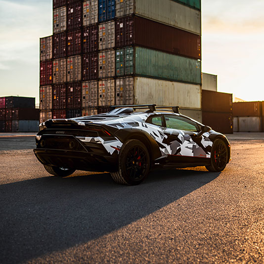 A white and black camouflaged sports car parked on pavement near stacked shipping containers at sunset.