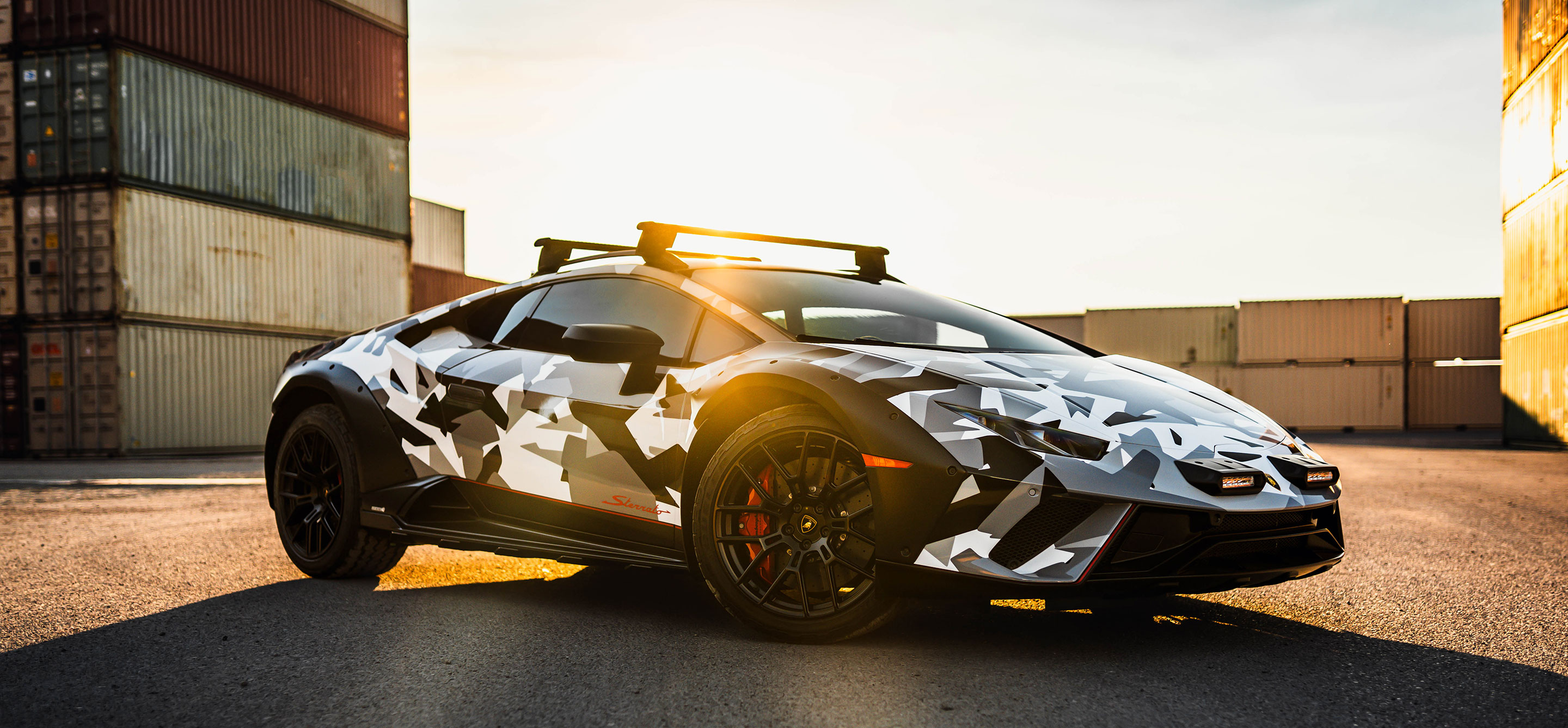 Camouflage-wrapped sports car parked near shipping containers at sunset. 