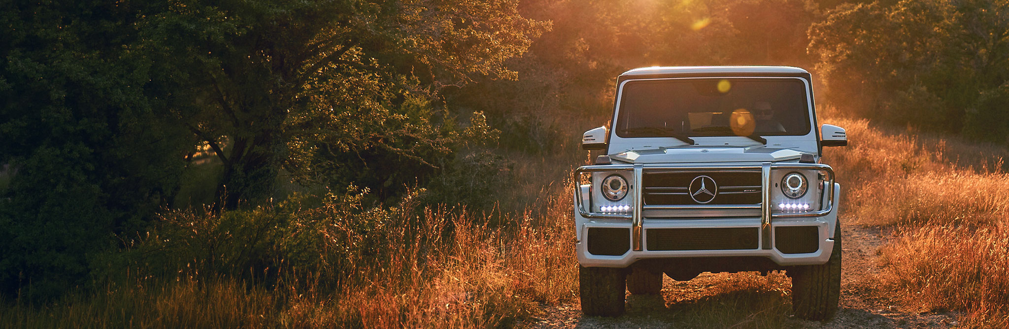 A white luxury off‑road SUV driving on a dirt trail at sunset, highlighting its rugged design, LED headlights, and premium Mercedes‑Benz styling. 