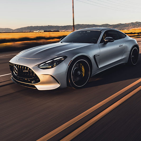 A silver sports coupe driving on an open road at sunset, with blurred pavement and fields in the background.