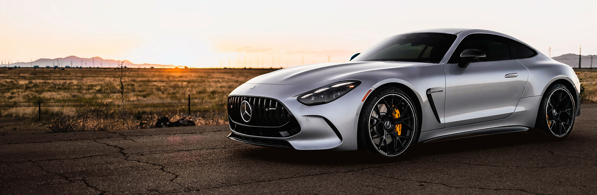 A silver sports coupe parked on a cracked desert road at sunset, with distant mountains silhouetted against the horizon. 