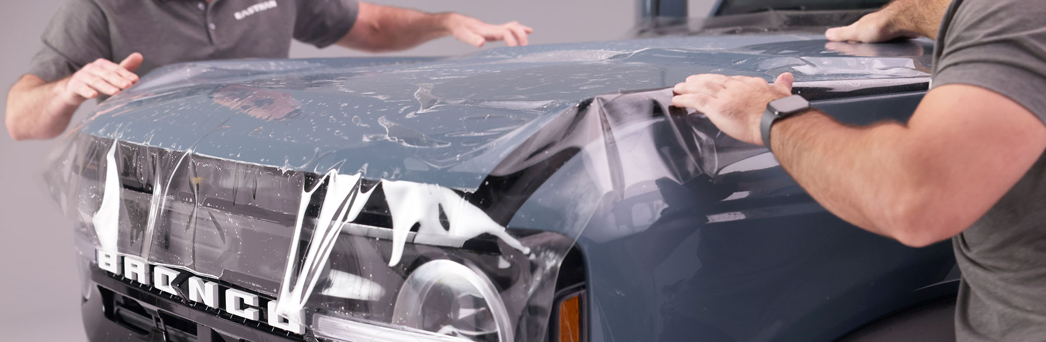 Two people applying protective film to the hood of a Bronco vehicle in a workshop. 