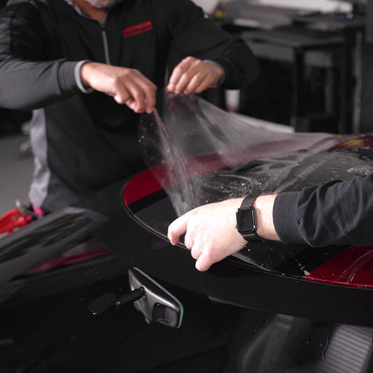 Two installers carefully apply a sheet of clear paint protection film to the rear section of a car.