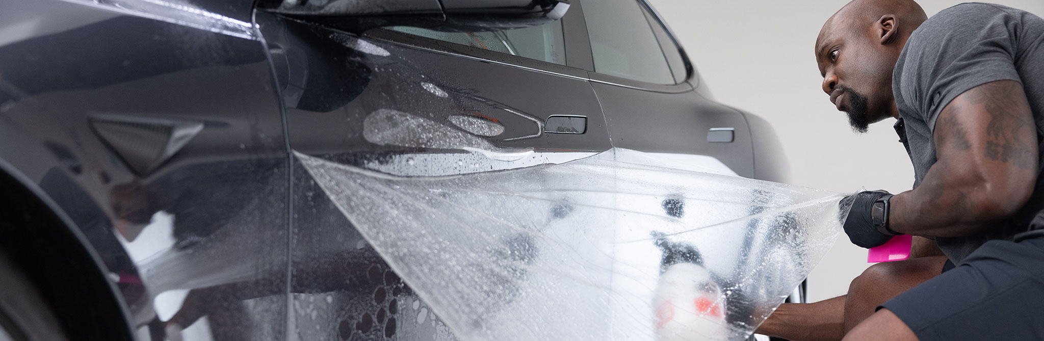 Person applying protective film to the side of a dark vehicle in a workshop. 