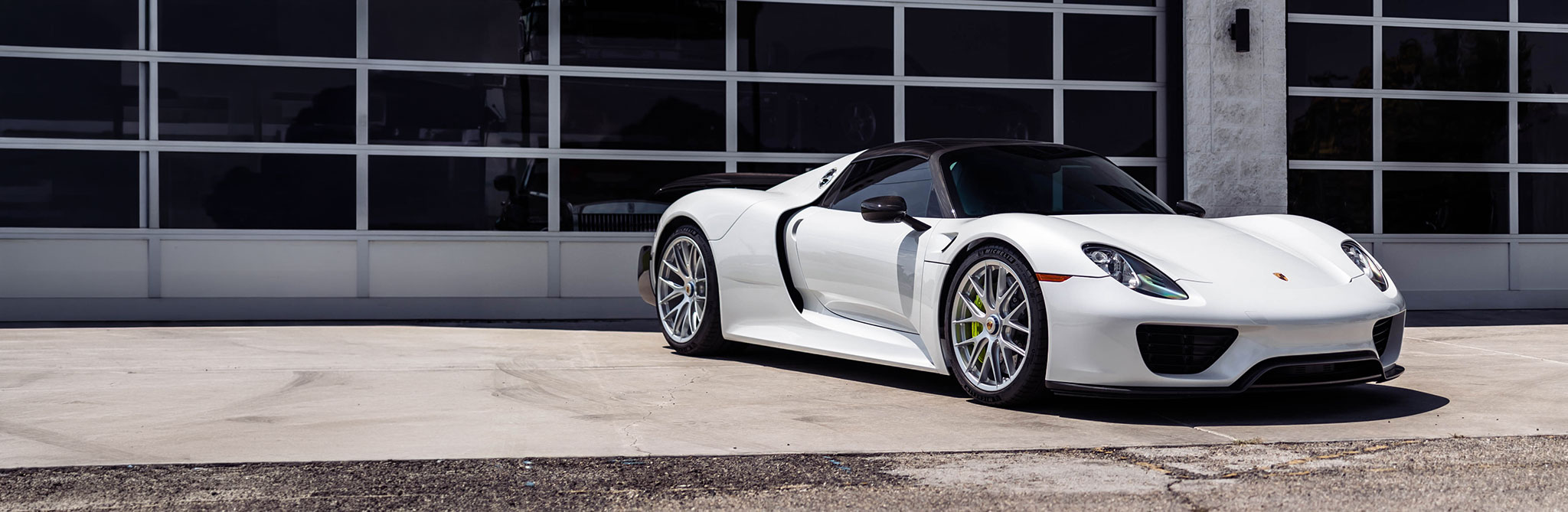 A white high‑performance sports car parked in front of a modern glass‑panel garage, shown from a front three‑quarter angle. 
