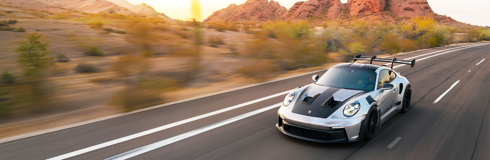 Silver and black sports car driving at high speed on a desert highway with red rock formations in the background. 