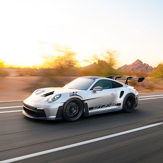 Silver high‑performance sports car driving on a desert highway at sunrise with mountains and desert brush in the background.