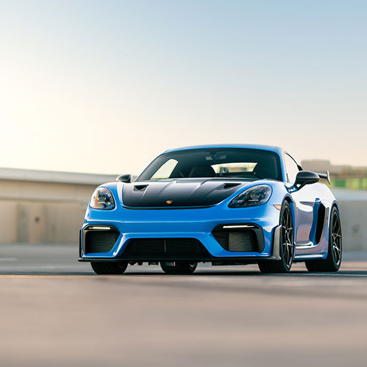 A blue sports car with a black hood is parked on an open rooftop lot at sunset.