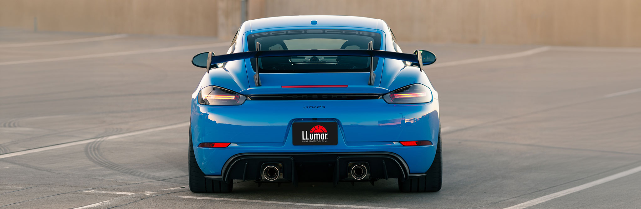 The rear view of a blue sports car with a large spoiler and dual exhausts, parked on a rooftop lot. 