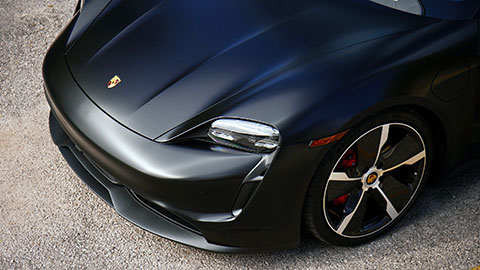 A close‑up top view of a matte black sports car, highlighting the front hood, headlights, and a large alloy wheel.
