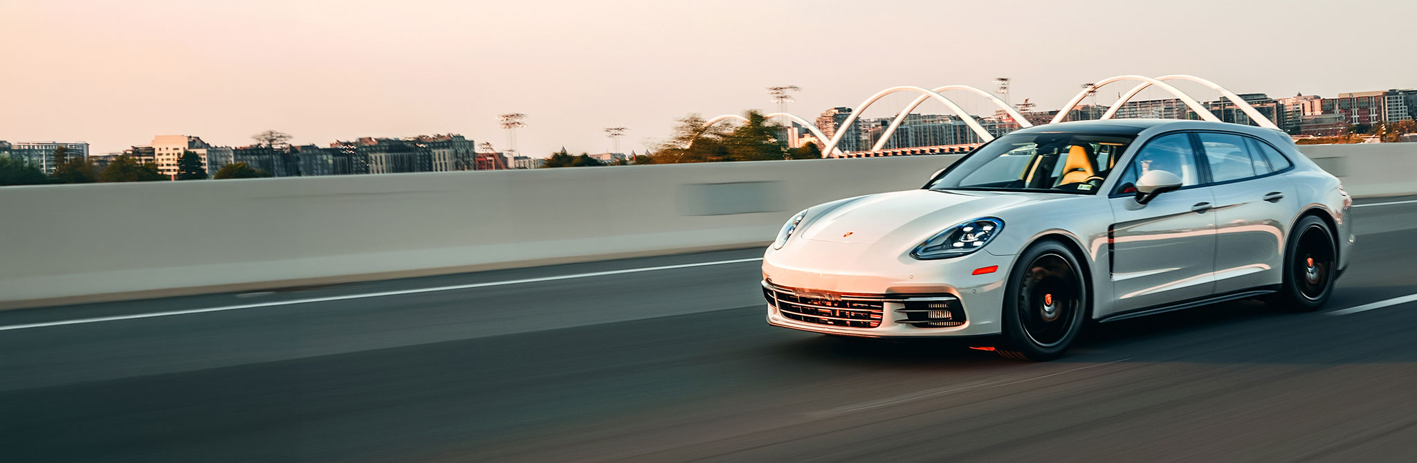 A white performance wagon driving on a city bridge at sunset with buildings and an arched structure in the distance. 
