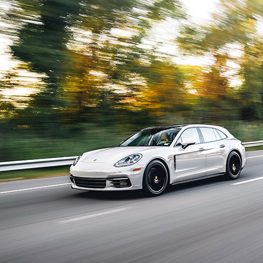 A white performance wagon driving on a highway with blurred autumn trees in the background.