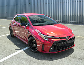 A red hatchback parked in an open lot with white parking lines and a metal-paneled building in the background.