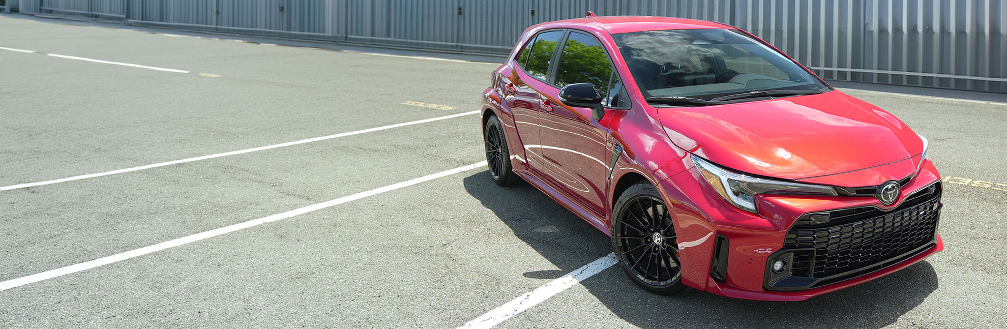 A red hatchback parked in an open lot with white parking lines and a metal-paneled building in the background. 
