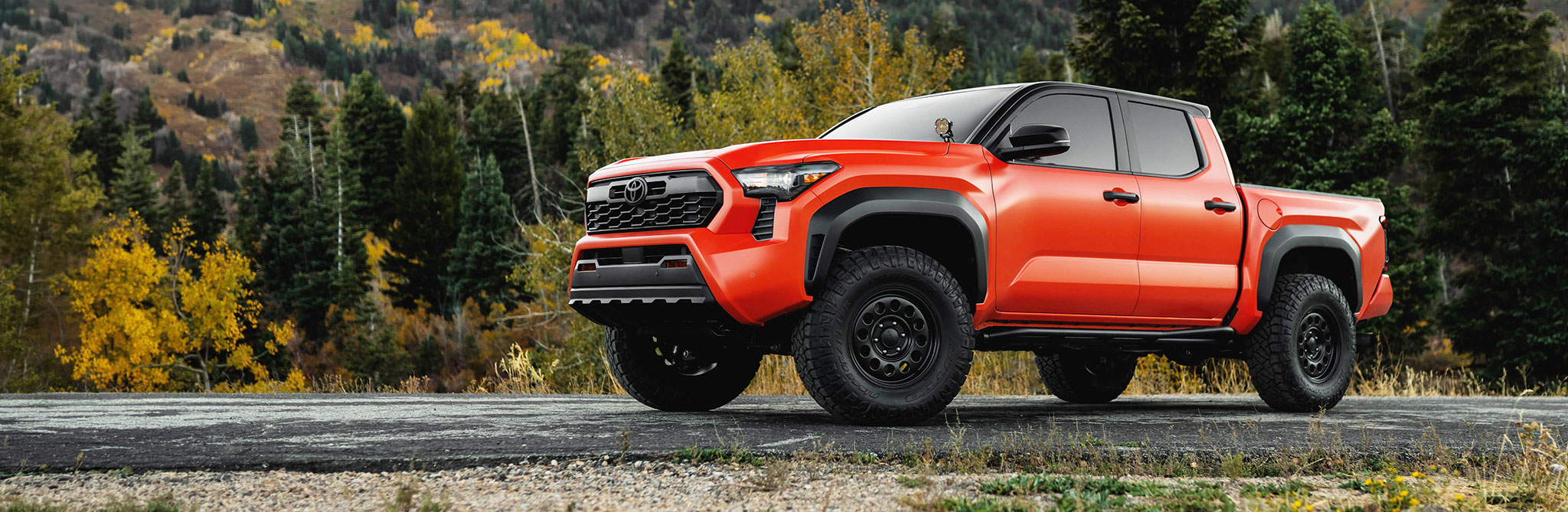 An orange off‑road pickup truck parked on a paved roadside with a backdrop of dense green and yellow autumn trees and mountain slopes. 