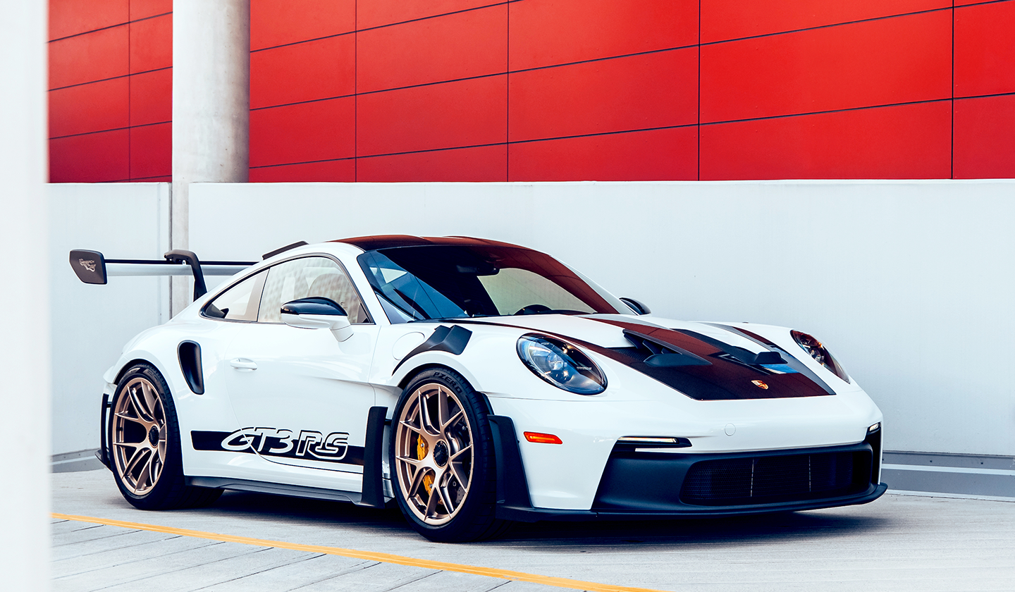 A white sports car with black accents and a large rear wing is parked on a modern concrete surface in front of a red and white building facade. 
