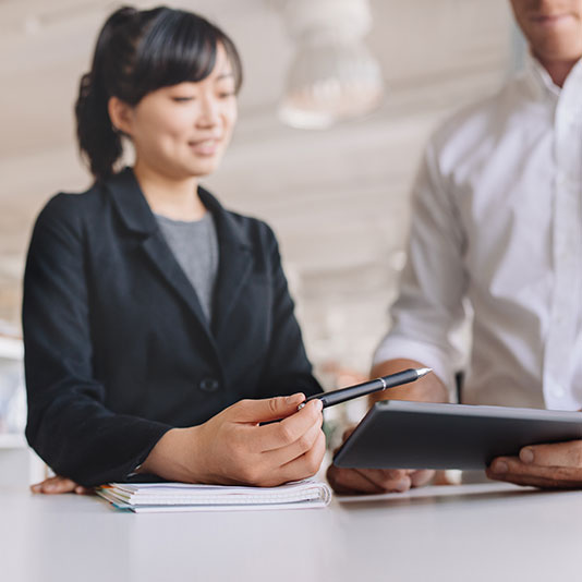 “A person in a dark blazer holds a pen over a notebook while another person beside them uses a tablet, both standing at a desk in a bright, modern workspace.