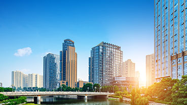 A city skyline featuring tall modern office buildings, residential towers, and a bridge over water, shown at sunrise with warm light reflecting off the structures.
