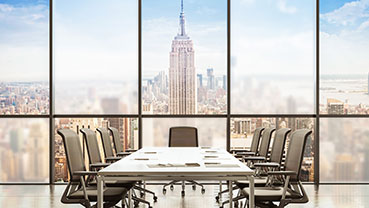 Large conference room featuring a long table and high‑back chairs, positioned in front of floor‑to‑ceiling windows showing a city skyline with tall buildings.