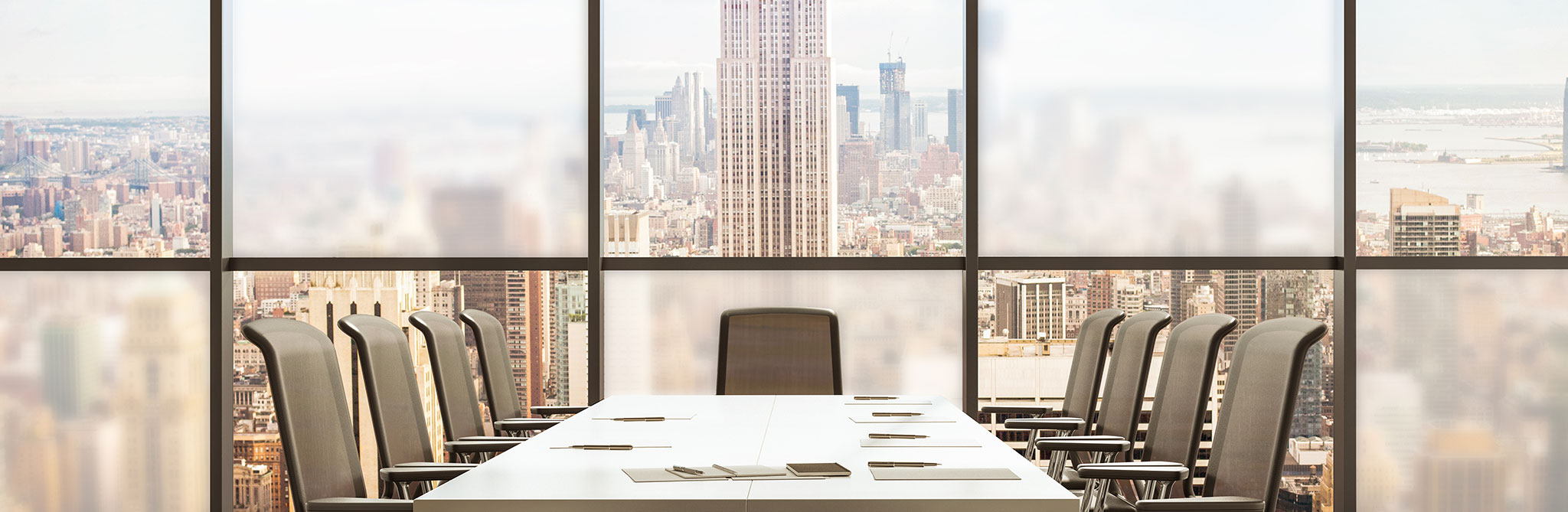 Large conference room featuring a long table and high‑back chairs, positioned in front of floor‑to‑ceiling windows showing a city skyline with tall buildings. 