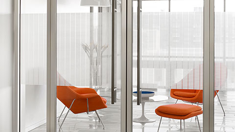 Modern office lounge with orange upholstered chairs and small white tables, viewed through glass walls with subtle horizontal privacy lines.