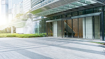 A modern commercial building entrance with floor‑to‑ceiling glass windows, metal framing, landscaped greenery, and a large overhang, with sunlight illuminating the exterior.