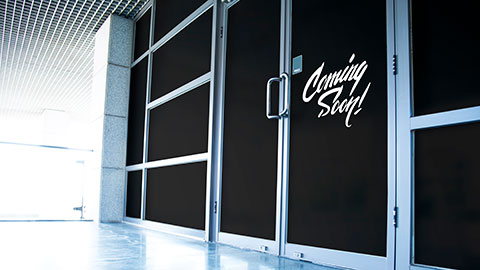 Glass storefront covered with black opaque film displaying the text ‘Coming Soon!’ in white script lettering near the door handle.