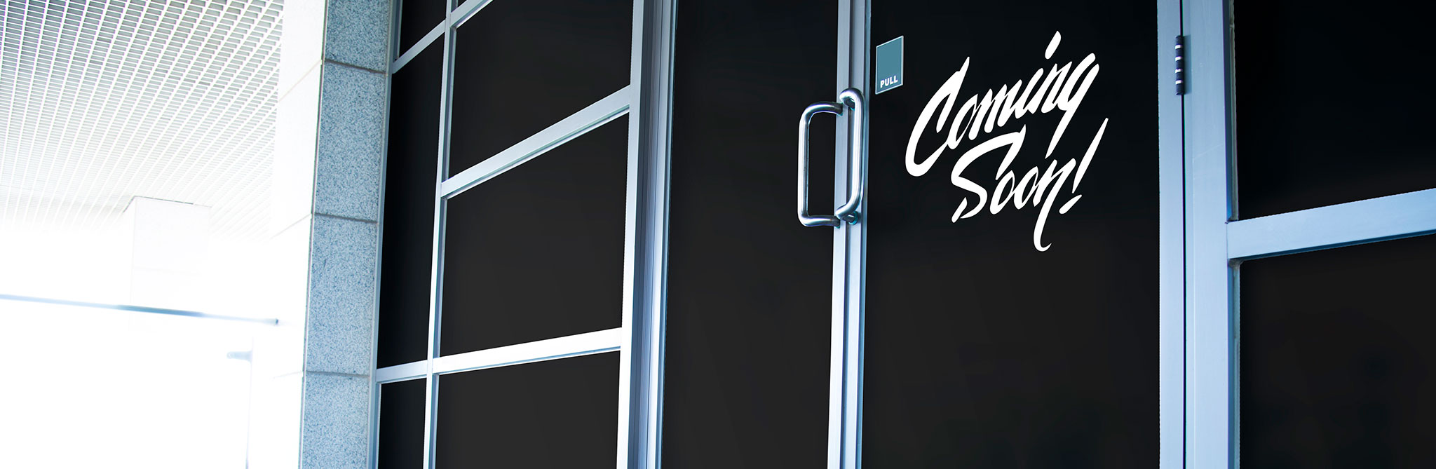 Glass storefront covered with black opaque film displaying the text ‘Coming Soon!’ in white script lettering near the door handle. 