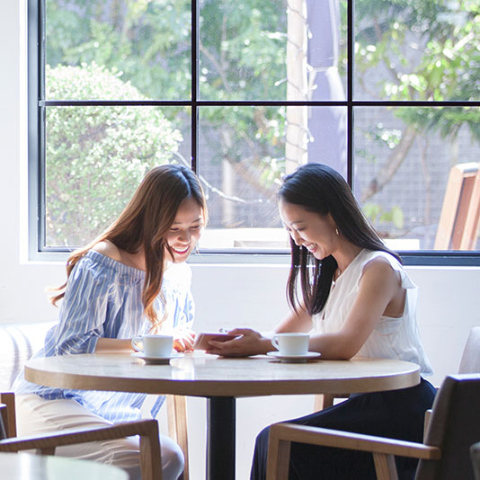 Two people sitting at a small round table with coffee cups in a bright café near a large window.