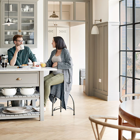 A modern kitchen with a gray island, glass‑front cabinets, and large windows, with two people seated at the island having breakfast.