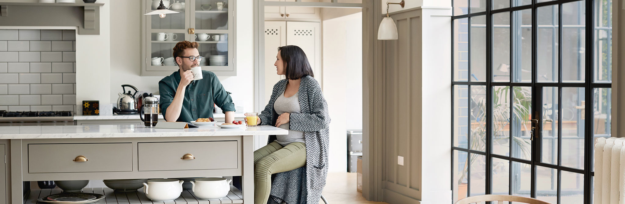 A modern kitchen with a gray island, glass‑front cabinets, and large windows, with two people seated at the island having breakfast. 
